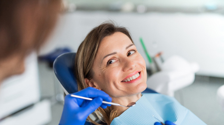 A smiling woman sitting in a dental chair, wearing a blue dental bib, while a dentist wearing blue gloves holds a dental mirror near her mouth. The background shows a clean, modern dental clinic setting.